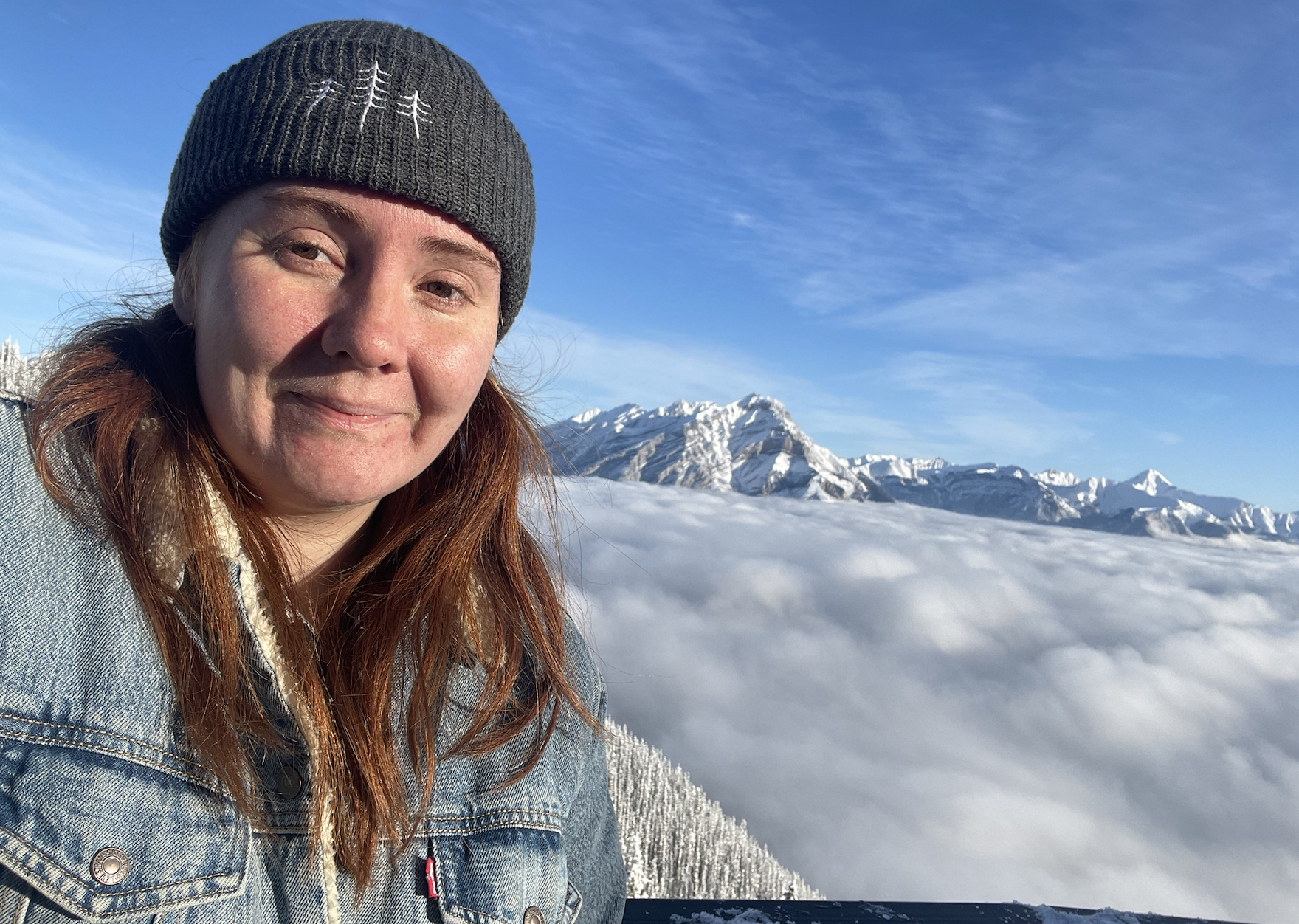 Emily wearing a winter hat and denim jacket and taking a selfie standing above the clouds on top of a mountain in Canada